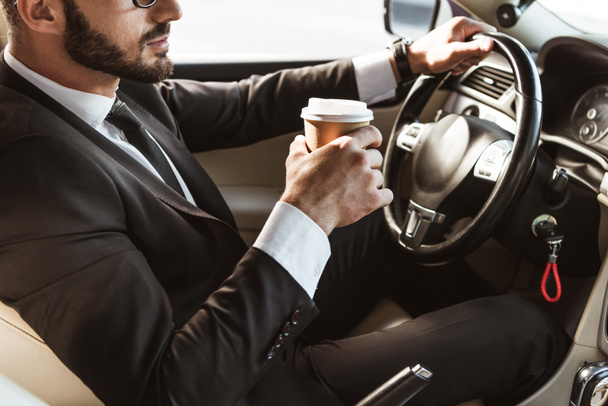 cropped image of driver in suit driving car and holding coffee in paper cup - Foto, afbeelding