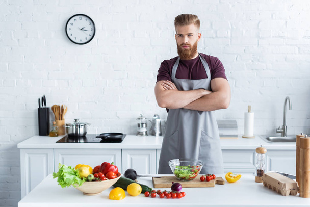 handsome bearded man in apron standing with crossed arms while cooking in kitchen - Φωτογραφία, εικόνα