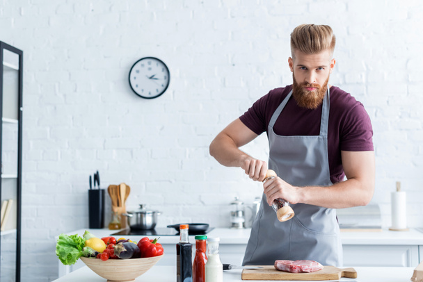 knappe bebaarde man in schort koken van heerlijke biefstuk en camera te kijken - Foto, afbeelding
