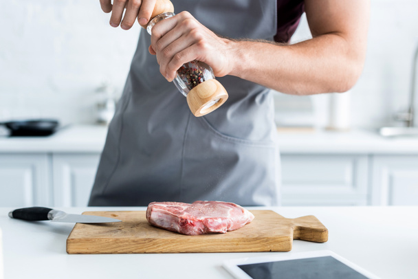oříznutý snímek muž v zástěře drží mlýnek na pepř při vaření hovězí steak - Fotografie, Obrázek