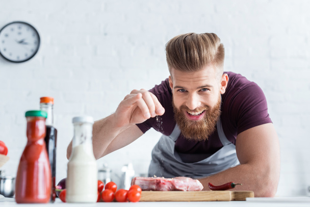 handsome bearded man in apron cooking delicious steak and smiling at camera  - Foto, Imagem