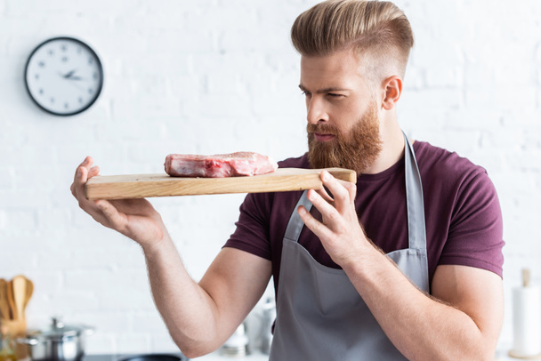 handsome bearded young man holding wooden cutting board with delicious raw beef steak - Foto, afbeelding