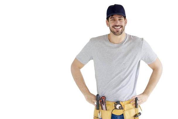 Portrait of happy young handyman with hands on his hip wearing tool belt and baseball cap while standing at isolated white background. - Фото, изображение