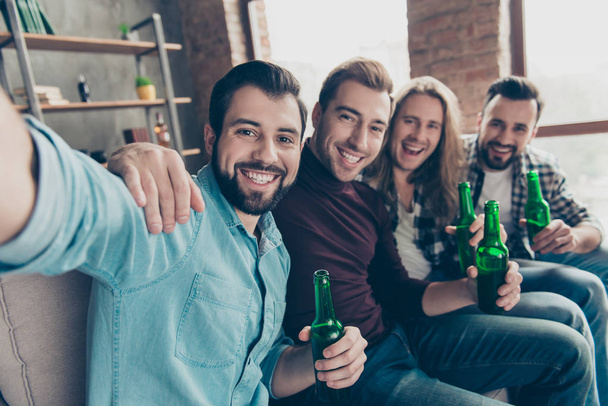 Self portrait of attractive, stylish, successful guys with modern hairstyle having bottles with lager in hands, shooting selfie while watching football match on tv, sitting in living room - Photo, image