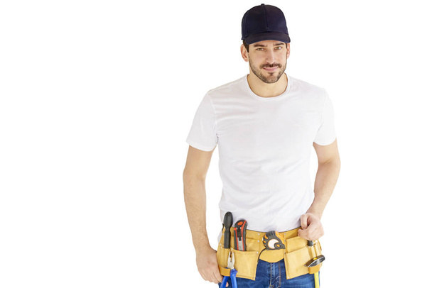 Portrait of handsome young handyman wearing baseball cap and tool belt while standing at isolated white background with copy space. - Fotoğraf, Görsel