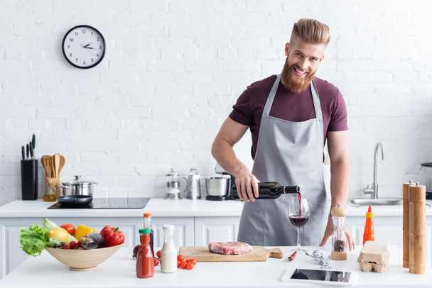 handsome bearded young man in apron pouring wine and smiling at camera while cooking in kitchen - 写真・画像