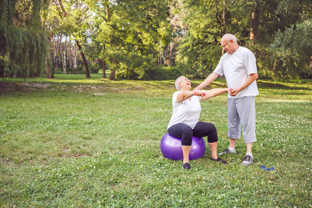 Concepto de estilo de vida saludable sonriente hombre y mujer maduros haciendo juntos ejercicios de fitness en la pelota de fitness en el parque
 - Foto, Imagen