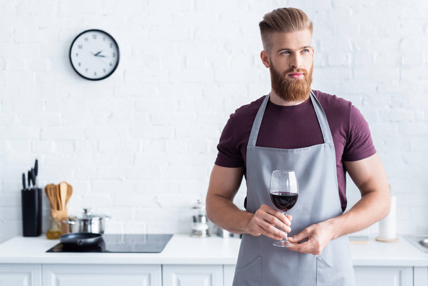 handsome bearded young man in apron holding glass of red wine and looking away in kitchen - Zdjęcie, obraz