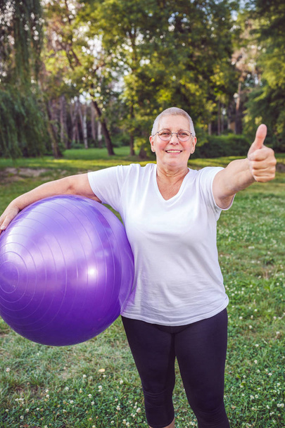 Daumen hoch für gesund trainierende lächelnde Seniorin mit Fitnessbällen im Park - Foto, Bild