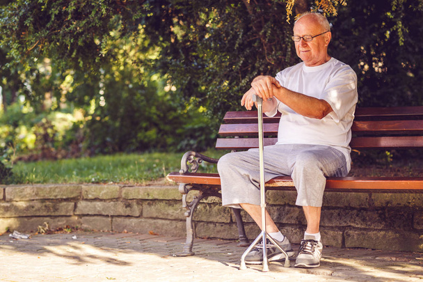 Triest oudere man met zijn wandelstok zittend op een bankje in het park - Foto, afbeelding