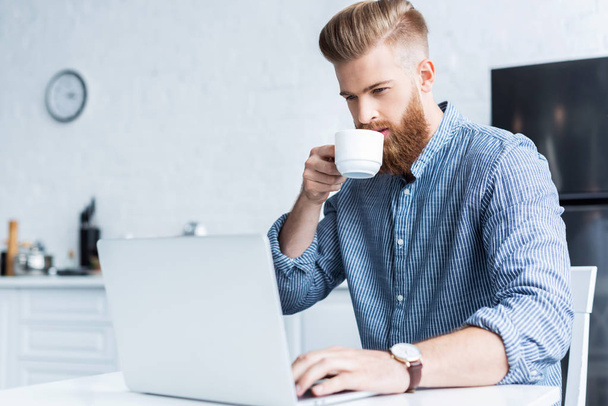 koffie drinken en het gebruik van laptop thuis knappe jongeman voor baard - Foto, afbeelding