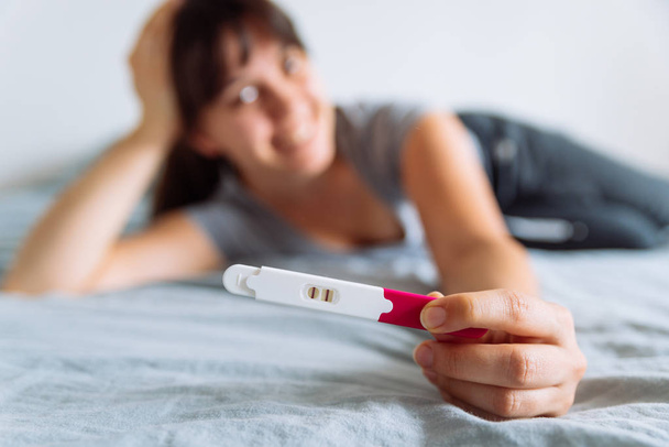 Genç mutlu kadın pozitif gebelik testi üzerinde seyir yatakta oturuyordu. arka plan bulanık - Fotoğraf, Görsel