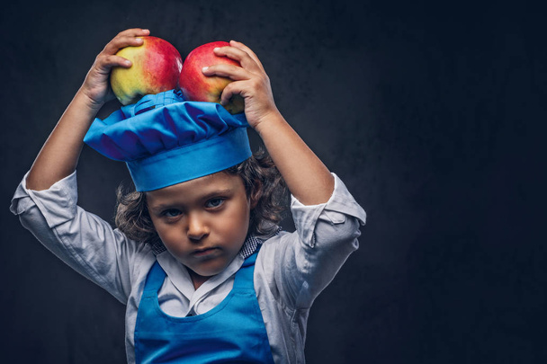 Portrait of a cute little boy with brown curly hair dressed in a blue cook uniform, has fun with apples at a studio. Isolated on the dark textured background. - Photo, Image