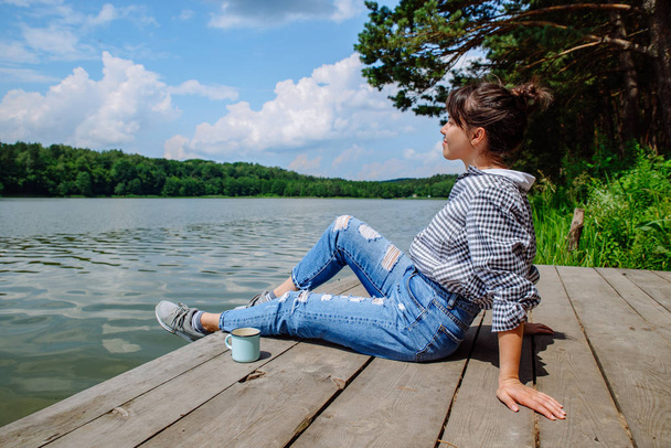 junge erwachsene Frau sitzt auf Holzsteg, trinkt Kaffee und blickt auf den See - Foto, Bild