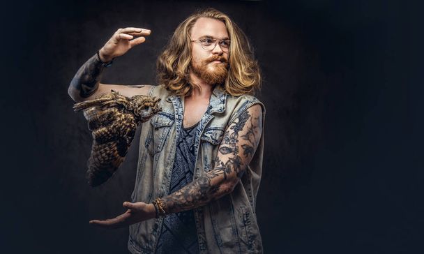 Retrato de un hombre pelirrojo hipster tatuado con el pelo largo y exuberante y barba llena vestido con una camiseta y chaqueta sostiene un mantiene el espantapájaros de un búho en un estudio. Aislado sobre un fondo oscuro
. - Foto, Imagen