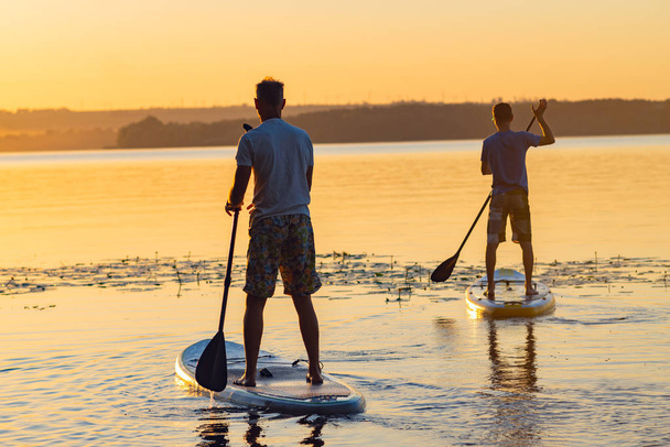 Men, friends sail on a SUP boards in a large river during sunrise. Stand up paddle boarding - awesome active recreation in nature. Back view.  - Photo, image