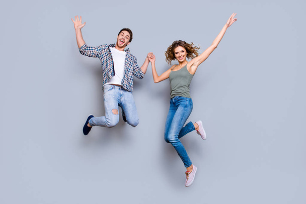Portrait of funky foolish couple in jeans sneakers jumping in air holding hands up enjoying time together isolated on grey background. Move motion concept - Zdjęcie, obraz