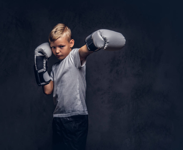 Školák boxer s blond vlasy, na sobě bílé tričko nosí Boxerské rukavice ukazuje kick boxu. Izolované na tmavém texturou pozadí. - Fotografie, Obrázek