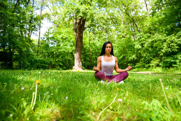 A beautiful young sporty girl sitting and meditating on the grass in the woods while wearing a leggings and a top - Photo, Image