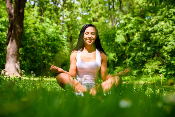 A beautiful young sporty girl sitting and meditating on the grass in the woods wearing a top and a short - Photo, Image