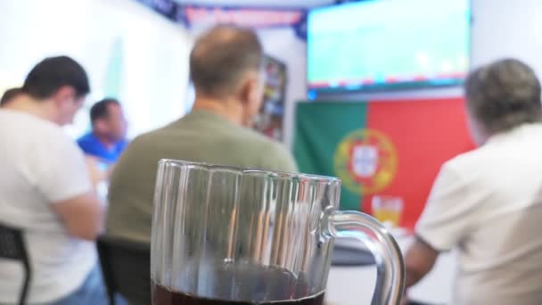 Kijken naar de voetbalwedstrijd op de televisie bij lokale pub. Voeding, bier. een mok met bier in focus, een Tv-scherm met een uitzending van een voetbalwedstrijd vervagen. 4k. - Video