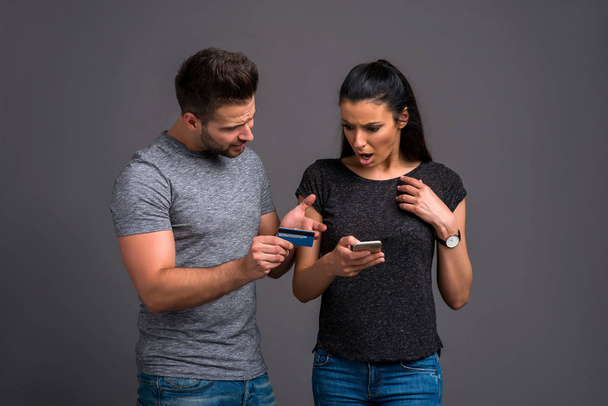 A handsome young guy showing his credit card to his beautiful girlfriend who is using a smartphone and they both feeling upset - Fotoğraf, Görsel