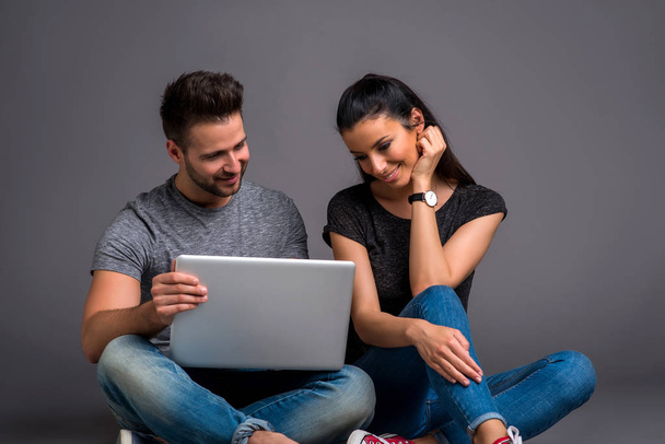 Ένα όμορφο νεαρό ζευγάρι κάθεται στο έδαφος και να βλέπουν ένα φορητό υπολογιστή και να χαμογελά - Φωτογραφία, εικόνα