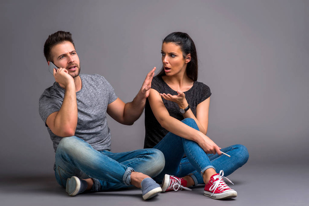 A handsome young guy sitting on the ground and ignoring his beautiful girlfriend while talking on his smartphone - Foto, imagen