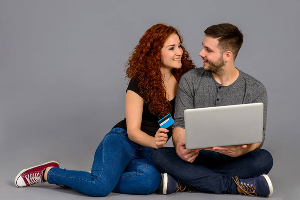 A handsome young guy holding a laptop while his girlfriend showing a credit card and they both smiling and sitting on the ground - Foto, Imagen