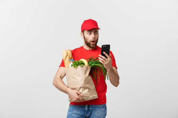 Liefermann hält Papiertüte mit Essen und Schock oder wütend über etwas in Handy auf weißem Hintergrund - Foto, Bild