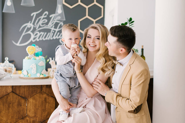 Beautiful young parents smile together with their one-year-old child at home in a beautiful interior in pastel colors with a festive blue cake. Family look. Happy birthday party - Foto, imagen