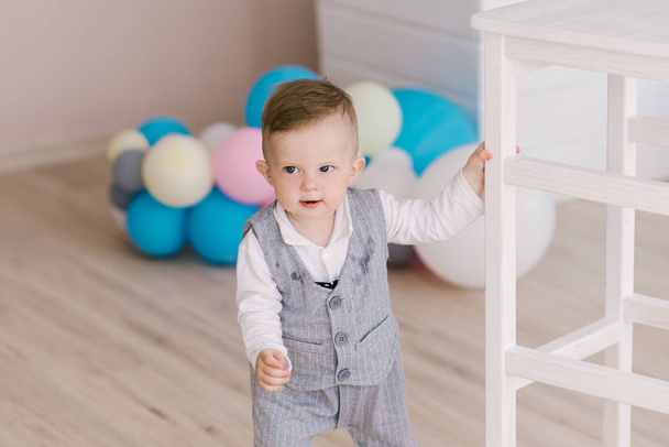 Happy cute boy in a gentleman's suit stands near the support on a light background, trying to get up, learning to walk. - Foto, Imagem