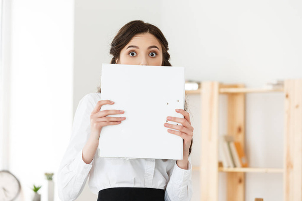 Young woman with surprised eyes peeking out from behind paper poster. Businesswoman holding big white banner in modern office. - Photo, Image