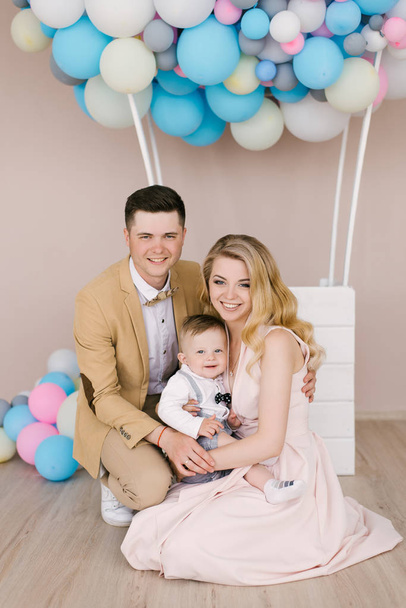 Beautiful young parents smile with their one-year-old child on pink and blue balloons background. Family look. Happy birthday party - Фото, зображення