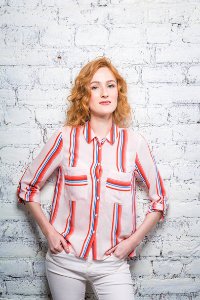 A beautiful young woman student with red curly hair and freckles is leaning against a brick wall of gray color. Dressed in a red striped shirt and white pants - Foto, Bild