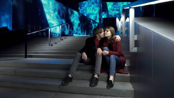 Νεαρό ζευγάρι κάθεται σε μια σκάλα μαζί σε έναν δρόμο το βράδυ. Φιλώντας ζευγάρι κάθεται στις σκάλες στο κέντρο της πόλης. Νεαρό ζευγάρι κάθεται σε σκαλοπάτια τη νύχτα. Όμορφο βράδυ για μια βόλτα γύρω από την πόλη - Φωτογραφία, εικόνα