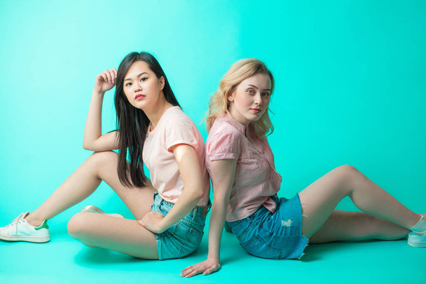 two multiethnic women sitting on the floor back to back isolated on blue background - Photo, image