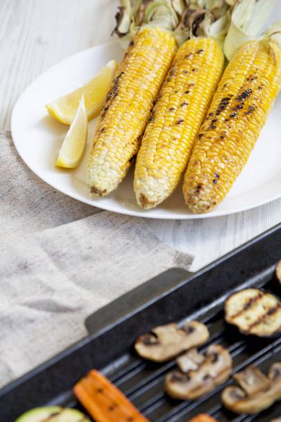 Grilled corn on a white plate, vegetables in a grilling pan, side view. Closeup. - Photo, Image