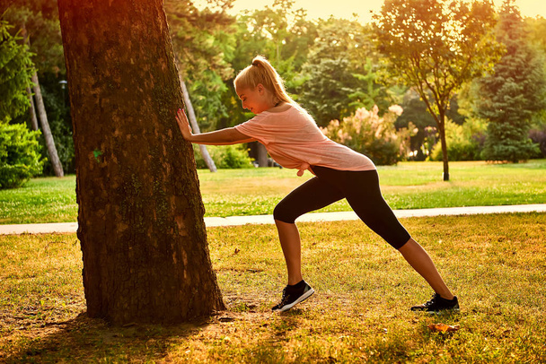 A beautiful young blonde woman leaning to a tree while stretching in a park in the sunset - Foto, Imagem