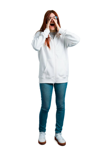 Young redhead girl in an urban white sweatshirt with glasses shouting with mouth wide open to the lateral and announcing something on isolated white background - Fotografie, Obrázek