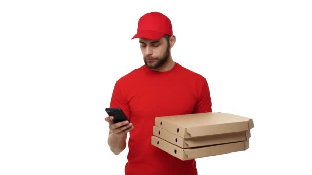 Delivery boy in a red uniform holding a stack of pizza boxes and checking order on mobile phone. Isolated over white background. - Footage, Video