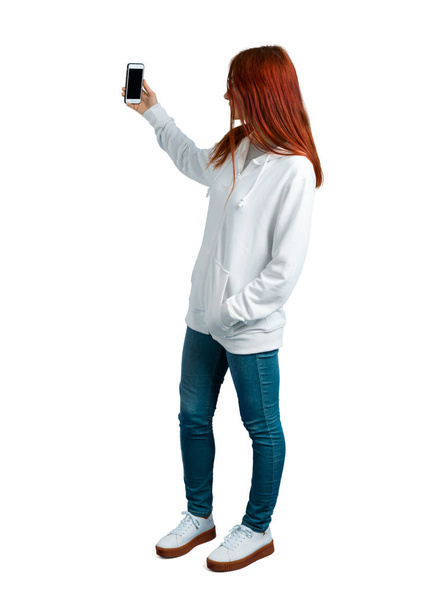 Mladá zrzka dívka v městské bílá mikina s brýlemi s selfie s mobilním telefonem - Fotografie, Obrázek