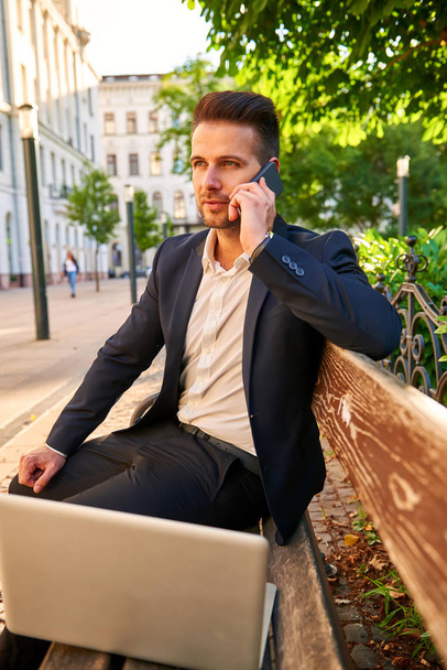 A handsome young businessman siting on a bench with his laptop on a sunny street next to a park while talking on his phone - Foto, immagini