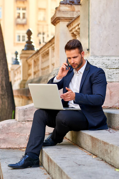 Красивый молодой бизнесмен сидит на лестнице на улице со своим ноутбуком и разговаривает по телефону.
 - Фото, изображение