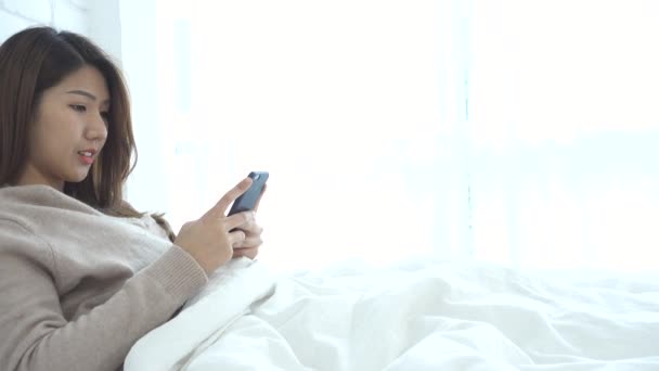 Happy Asian women are using smart phone on the bed in morning. Asian woman in bed checking social apps with smartphone. Smiling woman surfing net with cellphone at home. Mobile addict concept. - Video, Çekim