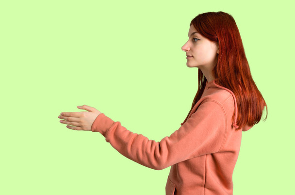 Young redhead girl with pink sweatshirt handshaking after good deal on green background - Foto, immagini