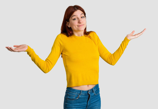 Young redhead girl with yellow sweater making unimportant and doubts gesture while lifting the shoulders and the palms of the hands on grey background - Foto, Bild
