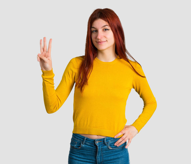 Young redhead girl with yellow sweater happy and counting three with fingers on grey background - Φωτογραφία, εικόνα