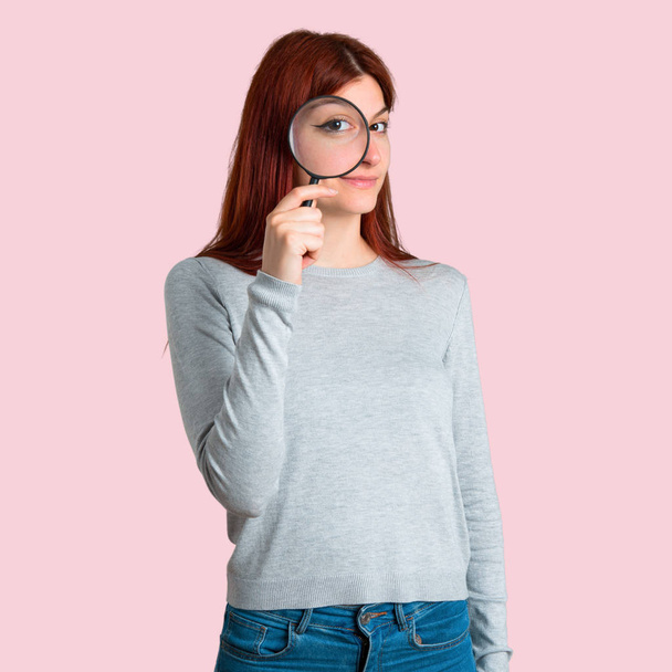 Young redhead girl taking a magnifying glass and looking through it on isolated pink background - Photo, image