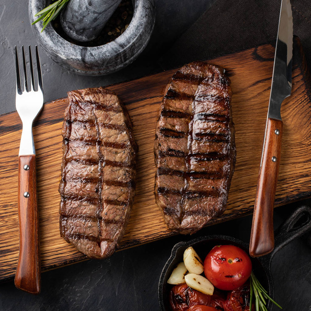 Closeup ready to eat steak Top Blade beef breeds of black Angus with grill tomato, garlic and on a wooden Board. The finished dish for dinner on a dark stone background. Top view - Foto, Imagem